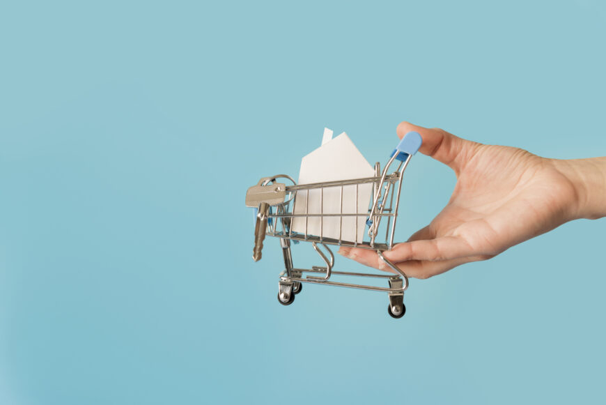 close up hand holding miniature shopping cart with paper house keys against blue background Как семьи с низким доходом могут оформить ипотеку